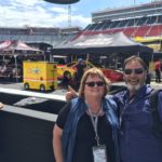 Two NASCAR fans smiling in the infield garage area at Bristol Motor Speedway with race cars and team haulers behind them.