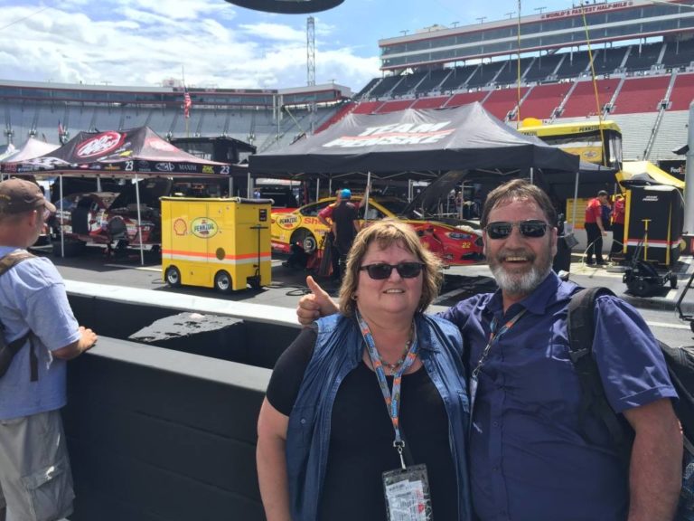 Two NASCAR fans smiling in the infield garage area at Bristol Motor Speedway with race cars and team haulers behind them.