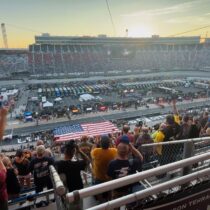 Packed grandstands at Bristol Motor Speedway during a NASCAR night race with fans and Bristol NASCAR ticket package guests standing for the national anthem as the American flag covers pit road.