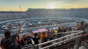 Packed grandstands at Bristol Motor Speedway during a NASCAR night race with fans and Bristol NASCAR ticket package guests standing for the national anthem as the American flag covers pit road.