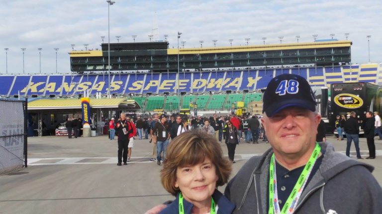 Fans smiling in front of the Kansas Speedway grandstands on race weekend, wearing NASCAR gear and credentials
