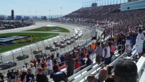 NASCAR Cup Series cars race past packed grandstands at Las Vegas Motor Speedway on a sunny day.