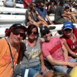 Group of NASCAR fans smiling in the grandstands during a sunny race day at Las Vegas Motor Speedway.