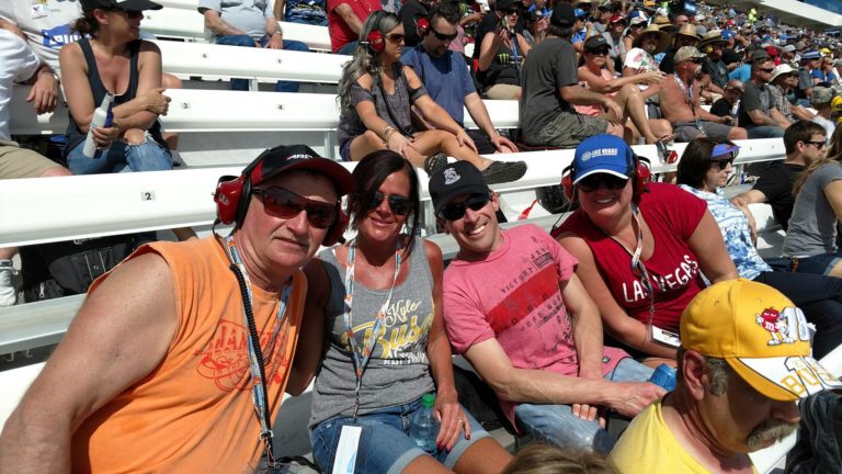Group of NASCAR fans smiling in the grandstands during a sunny race day at Las Vegas Motor Speedway.