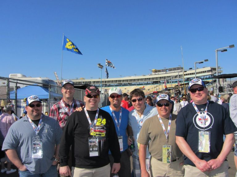 Group of RaceAway Phoenix NASCAR Ticket Package guests posing together at Phoenix Raceway with grandstands in the background.