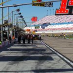 Pit road at Las Vegas Motor Speedway with NASCAR pit boxes and grandstands in the background on a sunny race day.
