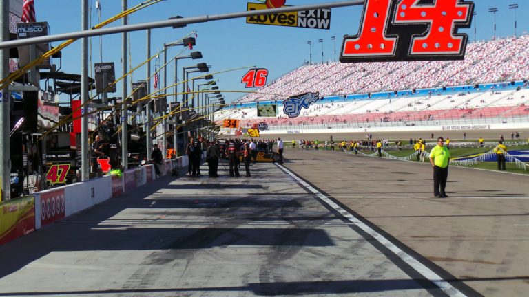 Pit road at Las Vegas Motor Speedway with NASCAR pit boxes and grandstands in the background on a sunny race day.