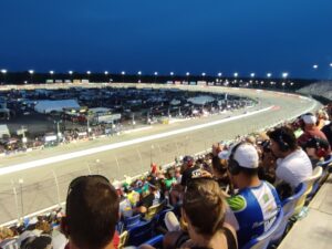 Fans in the grandstands cheer as cars roar under the lights at Darlington Raceway, part of the Southern 500 experience offered through RaceAway Darlington NASCAR Packages and Darlington NASCAR Tickets.