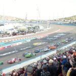 NASCAR Cup Series cars race past cheering fans in the grandstands at Phoenix Raceway.
