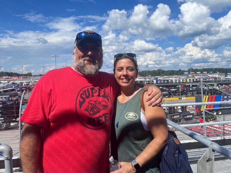 Smiling couple poses in the Darlington Raceway grandstands with the infield and pit road in the background.