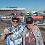 Two happy RaceAway guests on the track at Phoenix Raceway with grandstands and pit road in the background.