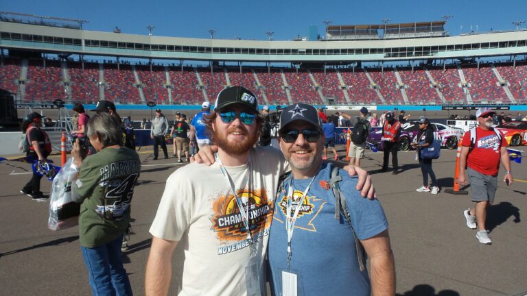 Two smiling NASCAR fans standing on the infield at Phoenix Raceway with grandstands behind them.