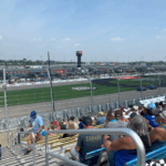Wide view of Iowa Speedway from the grandstands, with Iowa NASCAR Ticket Package guests watching cars on track and an American flag flying proudly overhead.