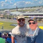 Two smiling NASCAR fans pose in the grandstands overlooking Talladega Superspeedway during race weekend.