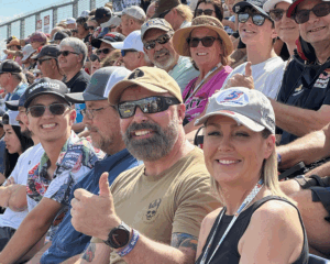 Smiling NASCAR fans seated in the grandstands at Talladega Superspeedway, with one fan giving a thumbs up.