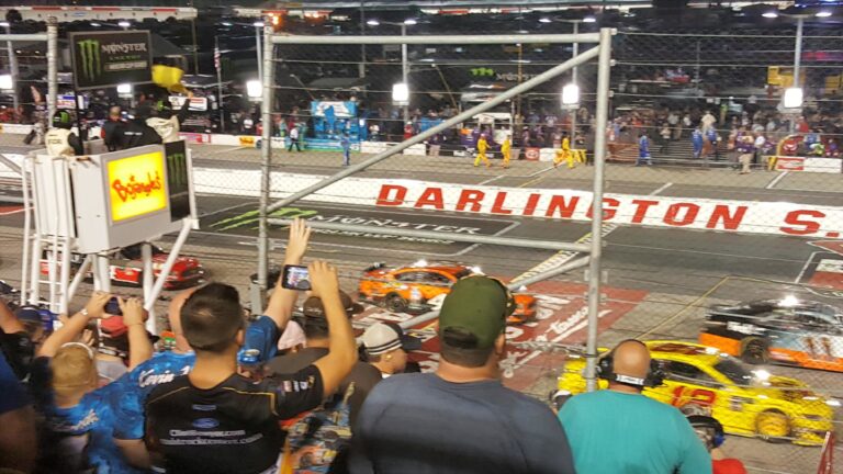 Fans cheer under the lights as stock cars roar past the start-finish line at Darlington Raceway during the Southern 500, part of the excitement offered in Darlington NASCAR Packages.