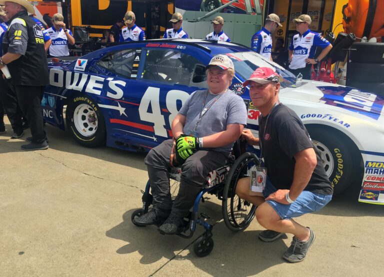 Two NASCAR fans, one in a wheelchair, posing in front of the No. 48 Lowe’s race car in the Charlotte Motor Speedway garage area.