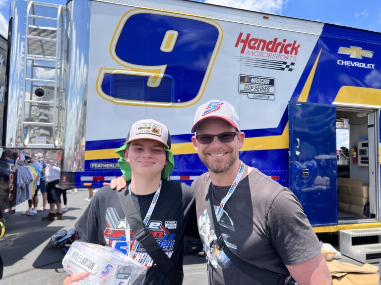 Father and son smiling in front of the Hendrick Motorsports hauler at Iowa Speedway during their RaceAway Hospitality weekend.