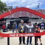 Three Talladega NASCAR package guests pose inside a red “This Is Talladega” frame at Talladega Superspeedway with grandstands full of spectators in the background.