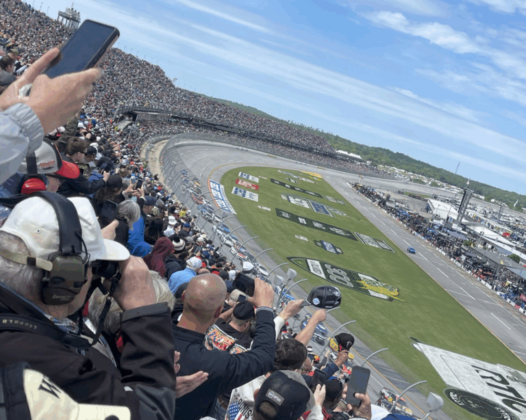 Crowd cheering as NASCAR Cup Series cars race past the grandstands at Talladega Superspeedway.