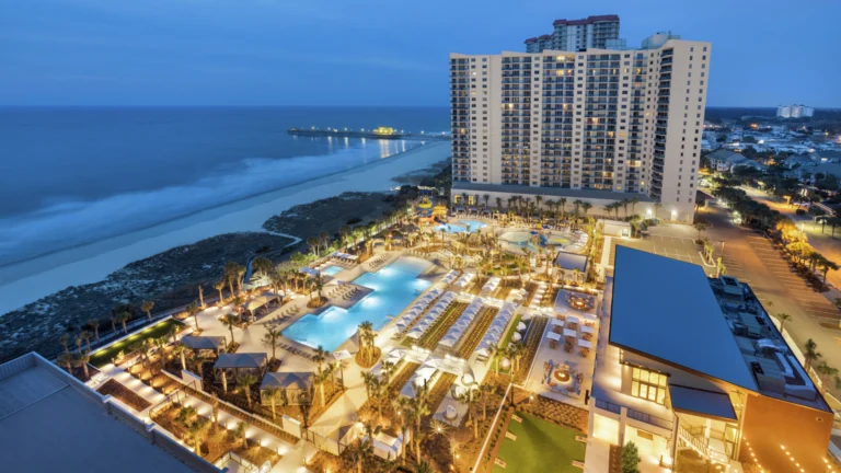 Evening aerial view of a Myrtle Beach resort with illuminated pools and oceanfront towers, included in Darlington NASCAR Packages.