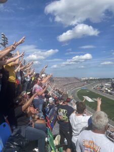 Fans raise their arms and cheer from the grandstands at Daytona International Speedway during the Daytona 500, with the frontstretch in view.
