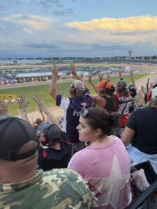 Race fans stand and raise their hands in the grandstands at Nashville Superspeedway during the 2026 Cracker Barrel 400 as the evening sky glows above the track.