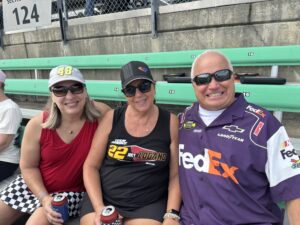 Three RaceAway guests smile from their seats at Kansas Speedway before the 2026 Hollywood Casino 400, wearing NASCAR driver and team apparel.
