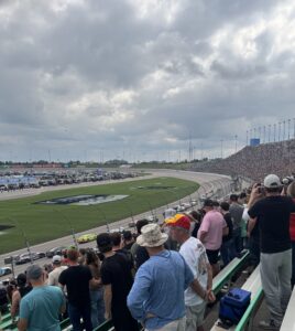 The NASCAR Cup Series field races past a packed grandstand at Kansas Speedway during the 2026 Hollywood Casino 400 under cloudy skies.