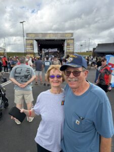 A pair of RaceAway guests pose near Charlotte Motor Speedway ahead of the October 2026 Bank of America 400, with the speedway grounds and an overcast Carolina sky setting the race-day scene.