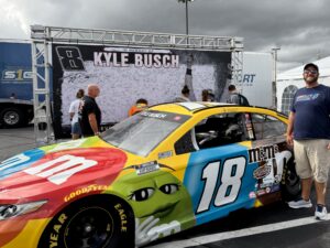 A RaceAway guest poses beside the No. 18 M&M’s Toyota and a Kyle Busch tribute display at Charlotte Motor Speedway during the 2026 Bank of America 400 weekend.