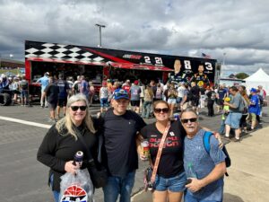 Four RaceAway guests pose in the fan zone at Charlotte Motor Speedway during the October 2026 Bank of America 400 weekend, with NASCAR merchandise haulers behind them.