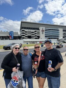 Four RaceAway guests enjoy the fan area at Charlotte Motor Speedway during the 2026 Bank of America 400 weekend, with team merchandise displays and race-day crowds in the background.