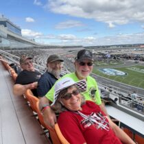 Four RaceAway guests gather for a race-day photo with Charlotte Motor Speedway rising behind them during the October 2026 Bank of America 400 weekend.