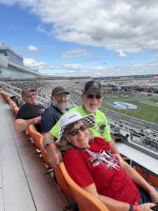 Four RaceAway guests gather for a race-day photo with Charlotte Motor Speedway rising behind them during the October 2026 Bank of America 400 weekend.